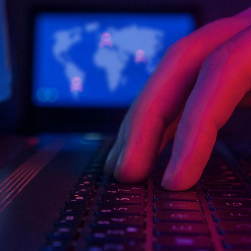 A cyber hacker rests their hand on a keyboard with code on the background screen.